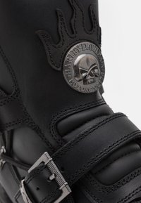 Bota de moto de cuero negro con una hebilla de metal, logotipo de calavera en relieve y detalle de diseño de llamas cerca de la parte superior.