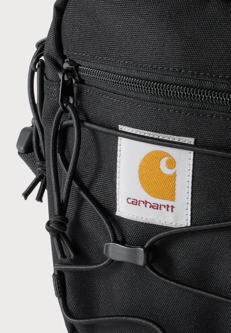 Close-up of black Carhartt bag with zipper and bungee cord detail, showing white label with yellow logo and red brand name.