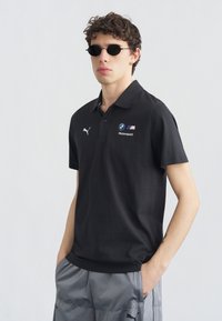 Νεαρός άνδρας που φοράει μαύρο πόλο BMW Motorsport και γκρι παντελόνι Puma, με στρογγυλά μαύρα γυαλιά ηλίου και σγουρά μαλλιά, στέκεται μπροστά σε γκρι φόντο.