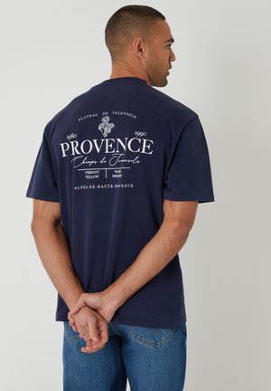 Homme aux cheveux courts portant un T-shirt bleu marine avec le texte blanc "Provence" et un motif floral, mains jointes dans le dos, de profil.