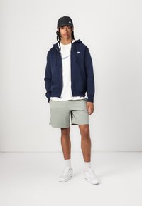 Sweatshirt azul-marinho com fecho de correr e bolsos, t-shirt branca com logótipo, calções verde-claro e ténis brancos. Boné preto com logótipo completa o visual.
