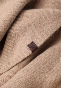 Bež villakangas, millel on ribitud tekstuur, millel on väike pruuni nahast silt, millel on tallatud karu logo.