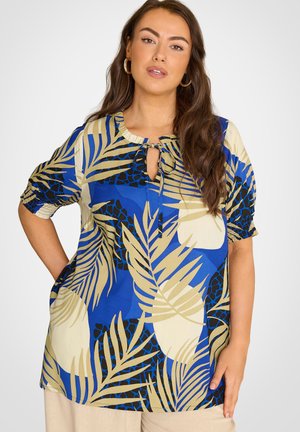 Frau mit langen braunen Haaren, die eine blau-beige Bluse mit tropischem Print und Schlüsselbund-Ausschnitt sowie beige Hosen trägt und mit der Hand in der Hüfte posiert.
