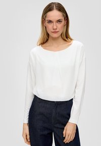Witte blouse met lange mouwen en een ronde hals, voorzien van een subtiele plooi aan de voorkant. Gepaard met donkerblauwe geklede broek. Gladde textuur.