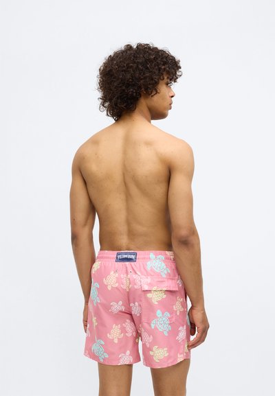 Shorts de baie roz dintr-un material cu imprimeu cu broaște țestoase în albastru deschis și galben. Talie elastică, cu un buzunar pe spate și eticheta cu un logo.