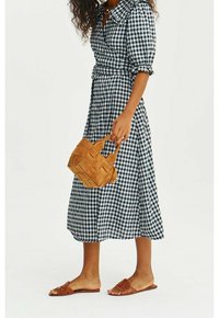 Femme portant une robe midi à carreaux noir et blanc, tenant un petit sac tressé beige et portant des sandales plates marron.