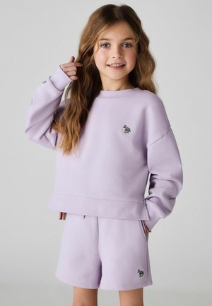 Junges Mädchen mit langen, welligen Haaren, das einen helllila Pullover und passende Shorts mit kleinen Zebra-Aufnähern trägt und in die Kamera lächelt.