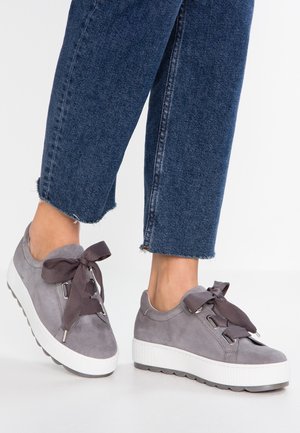 Feet wearing gray suede platform sneakers with large dark gray ribbons, paired with cropped blue denim jeans against a plain white background.