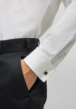 Witte shirt met een zwarte manchetknopen; de manchet is omgevouwen en heeft een vierkante metalen accent. Zwarte broek toont een soepele stoftextuur.