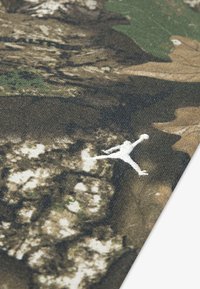 Maskovacia tkanina v rozličných zelených a hnedých odtieňoch, s bielou siluetou skáčúcej postavy. Mäkký, štruktúrovaný povrch.