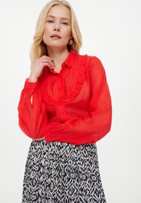 Rote, halbtransparente Bluse mit Rüschensaum, Knopfverschluss und langen Ärmeln. Kombiniert mit einem schwarz-weiß gemusterten Rock.