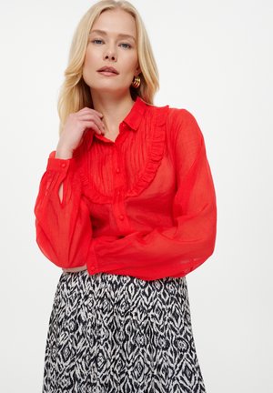 Rote, halbtransparente Bluse mit Rüschensaum, Knopfverschluss und langen Ärmeln. Kombiniert mit einem schwarz-weiß gemusterten Rock.
