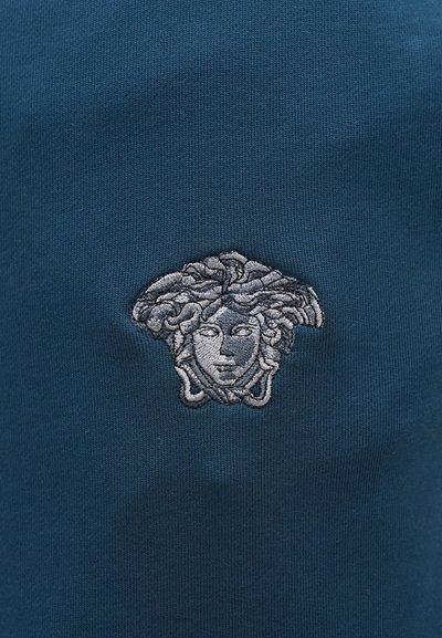 Tmavomodrá tkanina s detailným strieborným vyšívaným emblémom Medusy, zobrazujúcim prepracované vlasy a detaily tváre na hladkom povrchu.