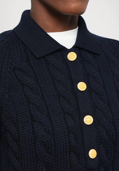 Polo en tricot torsadé bleu marine avec un col côtelé et quatre boutons jaunes sur le devant, mettant en valeur un design texturé et un contraste.