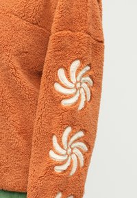 Oranžový plyšový pulovr s textúrovanou látkou. Obsahuje vyšívanie bieleho vírusu na rukávoch a predĺžený lem. Mäkký a útulný vzhľad.