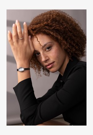 Ceas de mână argintie cu un cadran alb, purtat pe un brățară din piele maro, sprijinit pe o mână care zace pe obraz, alături de un inel simplu din argint.