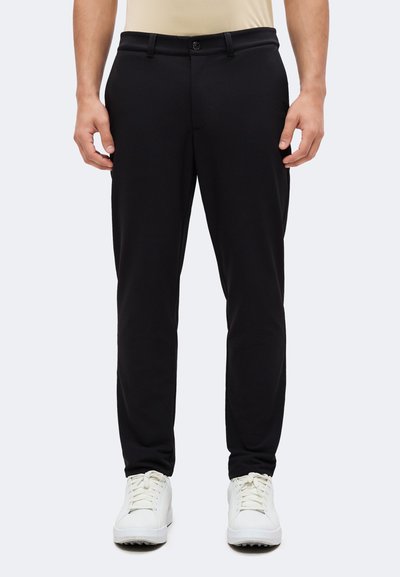 Homme portant un pantalon noir slim et des baskets blanches, debout avec les mains détendues le long du corps devant un fond clair uni.