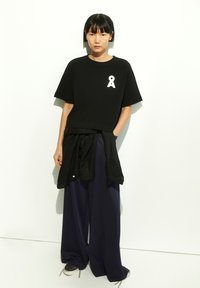 Black cotton oversized t-shirt features a white embroidered logo, paired with loose navy trousers and a black jacket tied at the waist.
