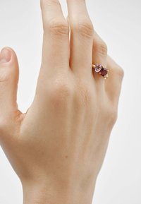 Anillo de oro rosado con dos piedras preciosas multifacéticas: un óvalo púrpura y un cuadrado rojo oscuro. Banda lisa y acabado pulido en el metal.