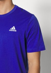 Sininen lyhythihainen t-paita, joka on valmistettu pehmeästä kankaasta, jossa on valkoinen Adidas-logo vasemmassa rinnassa ja klassinen pyöreä kaula-aukko.