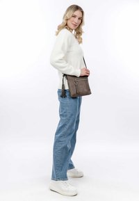 Brown crossbody bag made of soft material, featuring a front zipper pocket, tassel accent, and an adjustable strap. Pair with white sweater and jeans.