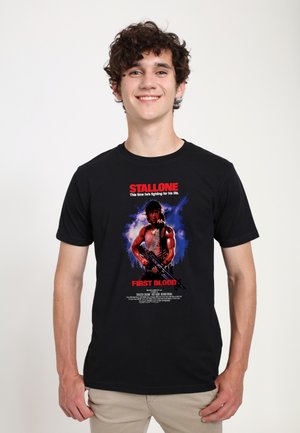 Ung mand med krøllet hår smiler iført en sort t-shirt med en bevæbnet muskuløs karakter og rød tekst fra filmen First Blood.