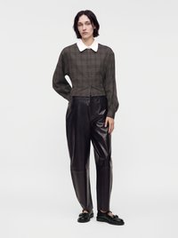 Femme aux cheveux courts et foncés portant une chemise à carreaux foncés boutonnée avec col blanc, un pantalon en cuir noir et des mocassins noirs, debout sur fond blanc.
