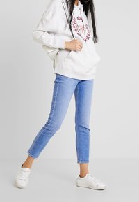 Žena nosí světle šedou mikinu s kapucí a leopardím vzorem, světle modré úzké džíny, bílé tenisky a malý bílý batoh, stojí před bílým pozadím.