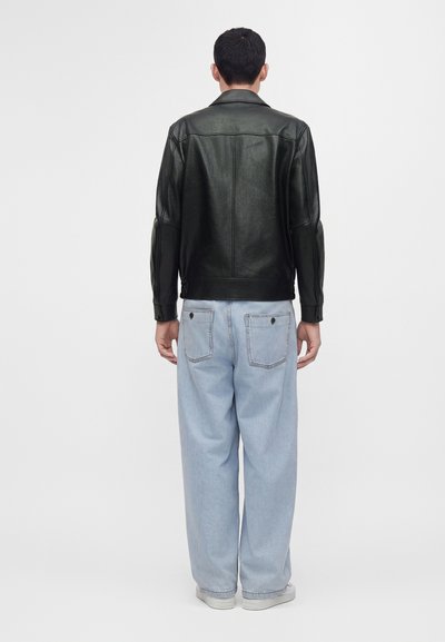 Man facing away wearing a black leather jacket, light blue wide-leg jeans with back pockets, and white sneakers on a plain white background.