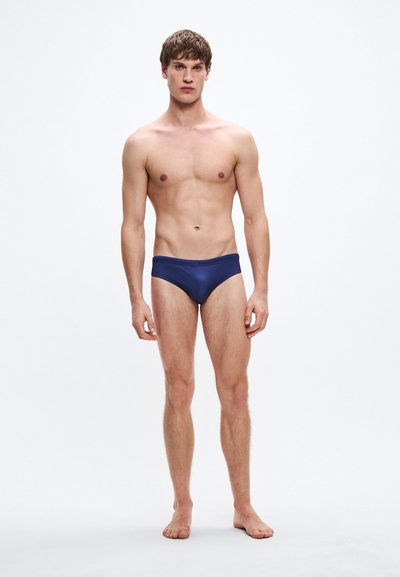 Cuecas de banho azul-marinho feitas de tecido suave, com design de cintura baixa e cintura elástica, usadas por um modelo masculino em pé.