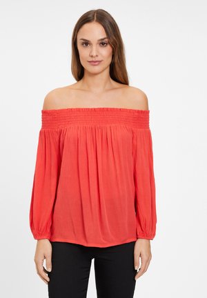 Top rouge décolleté avec encolure smockée, longues manches avec poignets élastiques et tissu texturé léger, associé à un pantalon noir.