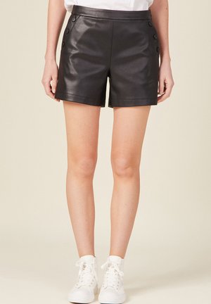 Zwarte leren shorts met een hoge tailleband, voorzien van knoopaccenten aan de voorkant en een gladde textuur. Gecombineerd met witte sneakers.