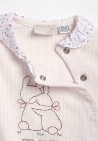 Body de bébé rose pâle avec col en dentelle florale, boutons-pression et contours brodés de deux lapins s'enlaçant.