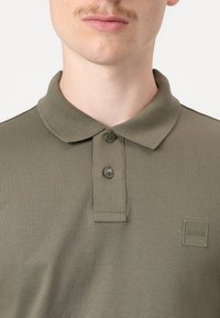 Olíva zöld pólóing, textúrált anyagból készült; gallérral, két gombos nyakkal és egy négyzet alakú logopatch-el a mellkason.