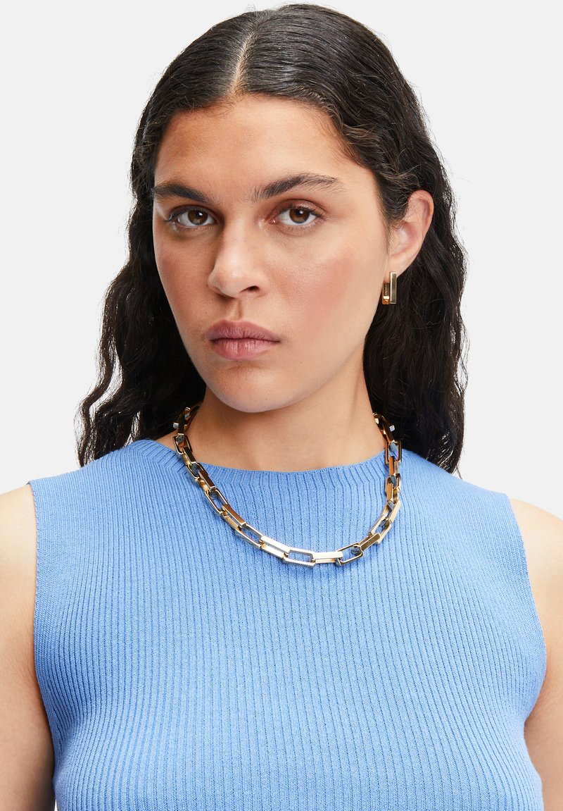 Collier en métal doré avec des maillons rectangulaires au fini texturé. Porté sur un haut sans manches côtelé bleu clair.