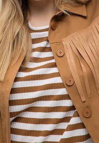 Giacca in suede marrone con dettagli di frange e due bottoni marroni, indossata sopra una maglietta a righe bianche e marroni con una texture a coste.