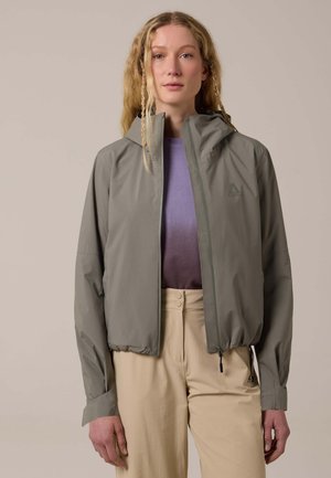 Woman with long, braided blonde hair wears a light gray zip-up jacket over a purple top and beige high-waisted pants against a plain background.