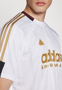 Vit idrottströja med vertikala ränder, guldaccents och svart ärmdetalj. Innehåller Adidas-logotyp i guld på bröstet.