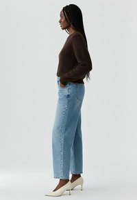 Pull en mélange marron, jeans droits bleu clair, et chaussures à talons blanches. Le mannequin se tient de profil devant un fond uni.