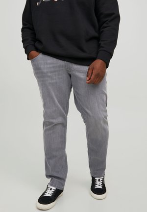 Osoba w jasnoszarych dżinsach, czarnych trampkach z białymi sznurowadłami oraz czarnej bluzie z rękami częściowo w kieszeniach.