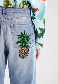 Jeans de mezclilla con un gran parche de piña de lentejuelas multicolores en el bolsillo trasero, con una camisa de patrón floral en azul claro visible.