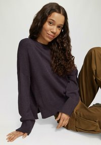Donkerpaars losgebreid sweater met ribgebreide manchetten en zoom, gecombineerd met bruine wijde broek; model zit, toont casual kleding en accessoires.