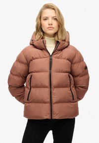 Chaqueta acolchada marrón con capucha, que presenta un cierre con cremallera al frente, dos bolsillos laterales y puños elásticos; diseño acolchado con un acabado suave.