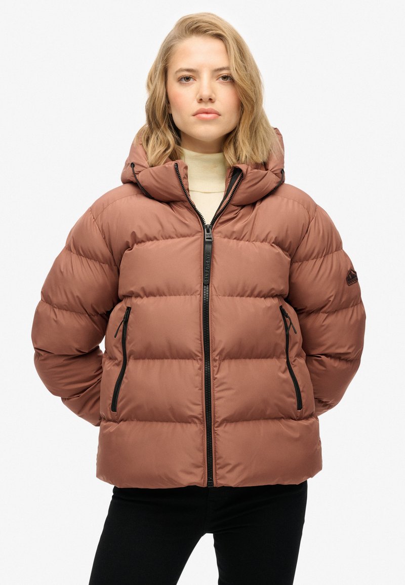 Chaqueta acolchada marrón con capucha, que presenta un cierre con cremallera al frente, dos bolsillos laterales y puños elásticos; diseño acolchado con un acabado suave.