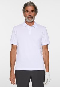 Vit kortärmad polo med klassisk krage, utrustad med en tre-knappsknappslå och en subtil logotyp på bröstet, i kombination med grå byxor.