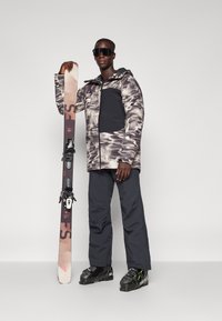 Chaqueta de esquí con un patrón en gris y negro, combinada con pantalones de esquí negros y botas de esquí negras. Sosteniendo una tabla de snowboard con fijaciones.