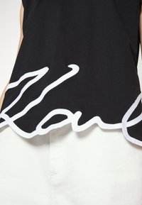 Camiseta negra con dobladillo en forma de ondas y detalle de texto bordado en blanco en la parte inferior. La tela parece suave con una textura lisa.