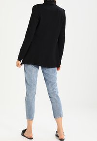 Blazer negro, corte entallado, hombros estructurados, tela suave, combinado con vaqueros azul claro y sandalias planas negras, visto desde atrás.