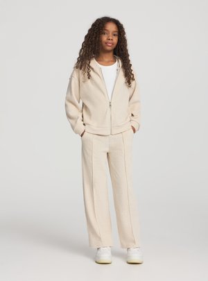 Giacca beige con zip e motivo testurizzato, abbinata a pantaloni a gamba larga coordinati e sneakers bianche. L'outfit è caratterizzato da un tessuto leggero e morbido.
