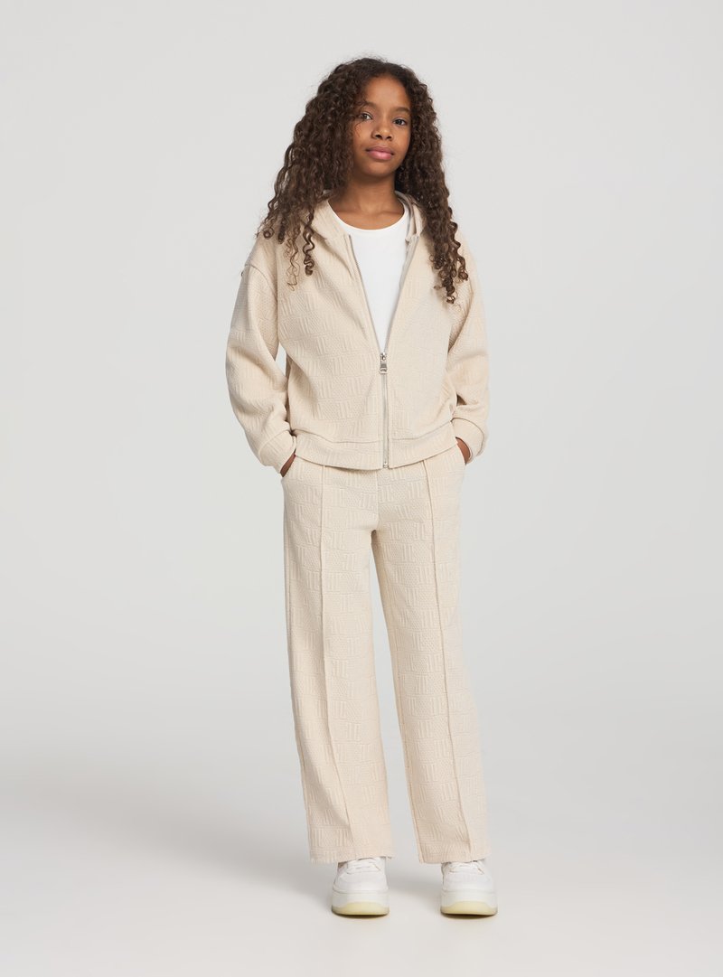 Giacca beige con zip e motivo testurizzato, abbinata a pantaloni a gamba larga coordinati e sneakers bianche. L'outfit è caratterizzato da un tessuto leggero e morbido.